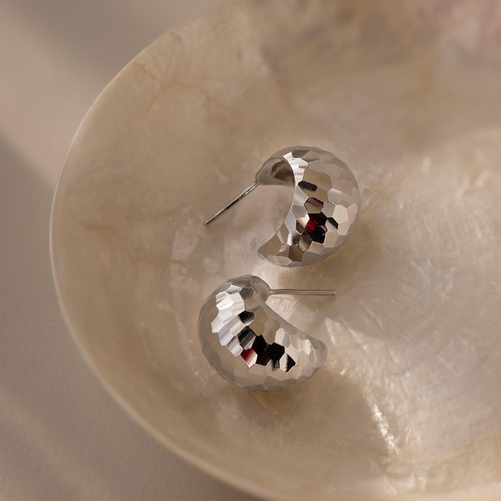 Bold Disco Earrings in hammered silver add retro glam, displayed on a round, textured beige dish—the perfect statement accessory.