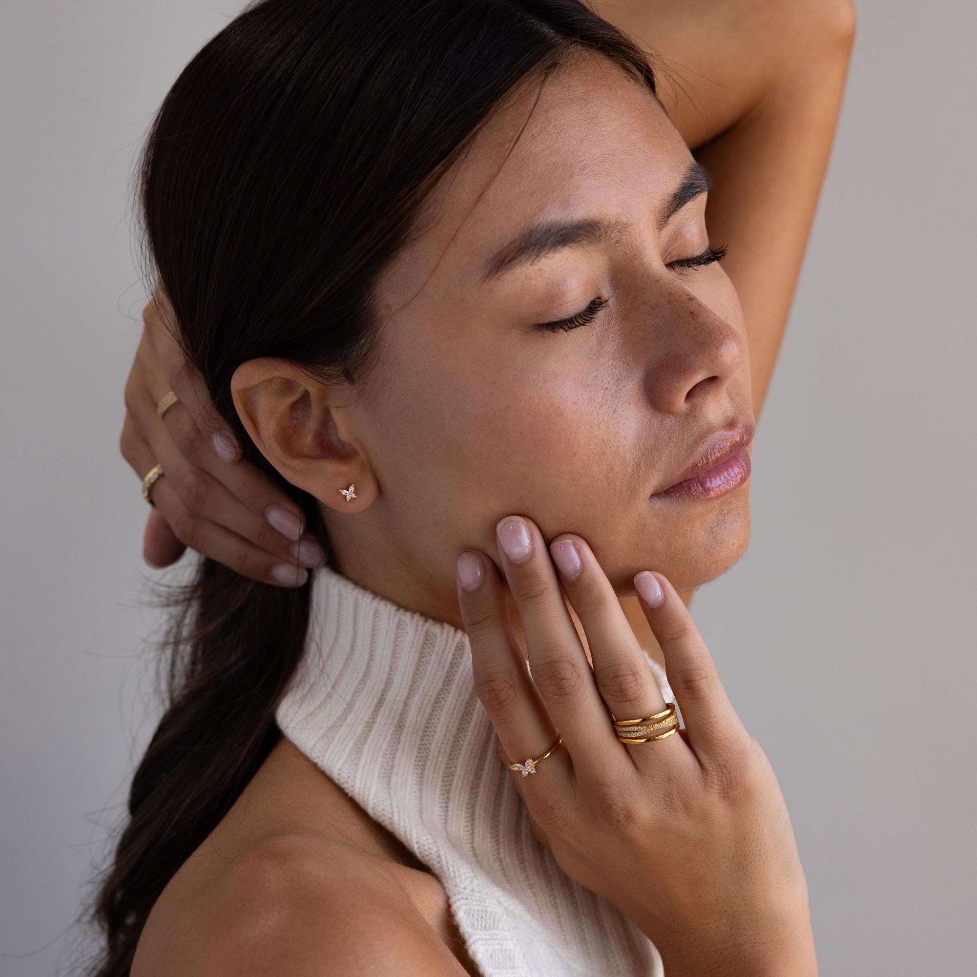 A woman with closed eyes wears gold jewelry, including delicate Butterfly Birthstone Flatback Studs, and touches her face and neck while dressed in a sleeveless cream turtleneck top.