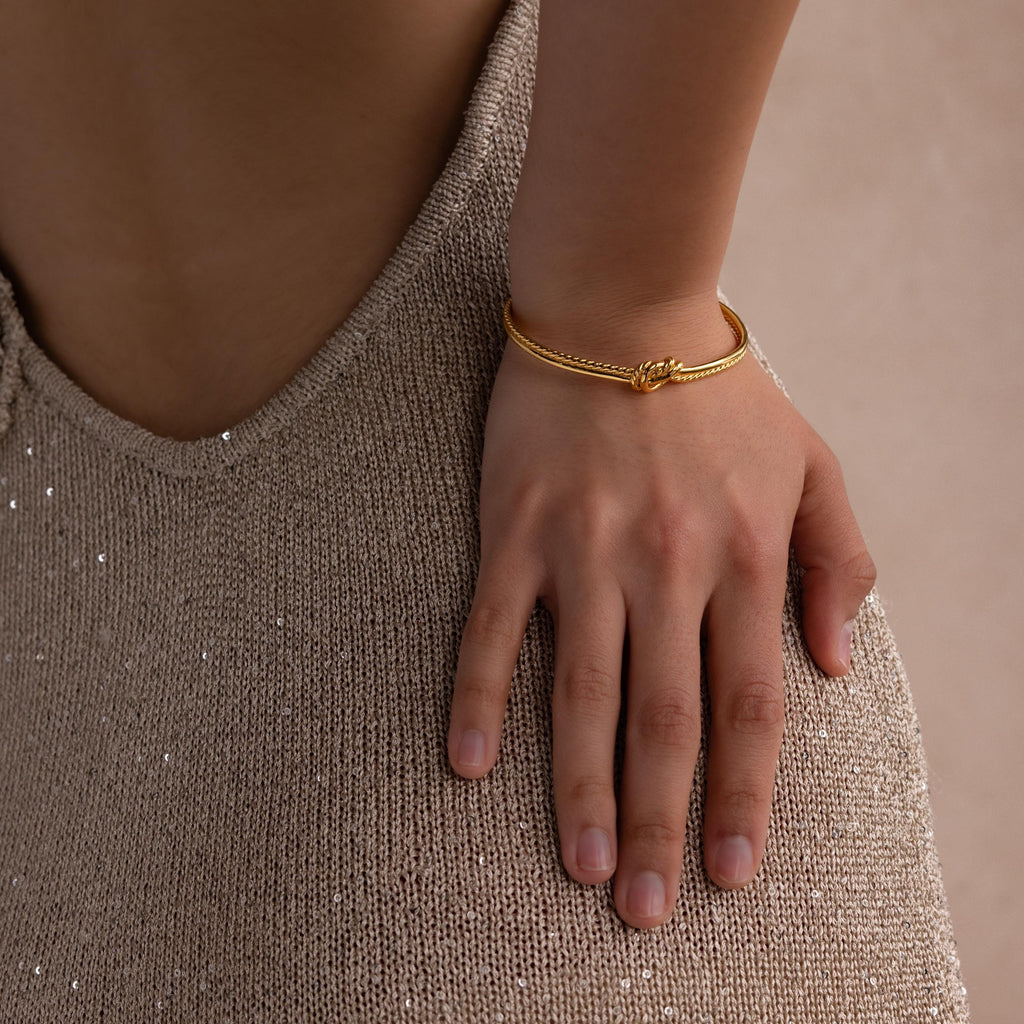 A hand rests on a sparkly beige skirt, showcasing the Duo Knot Bangle—a gold bracelet featuring a chic rope texture, perfect as an elegant gift.