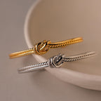 Two Duo Knot Bangles, one in gold and one in silver with a delicate rope texture, are displayed on a curved white ceramic dish—making them an elegant gift for any occasion.