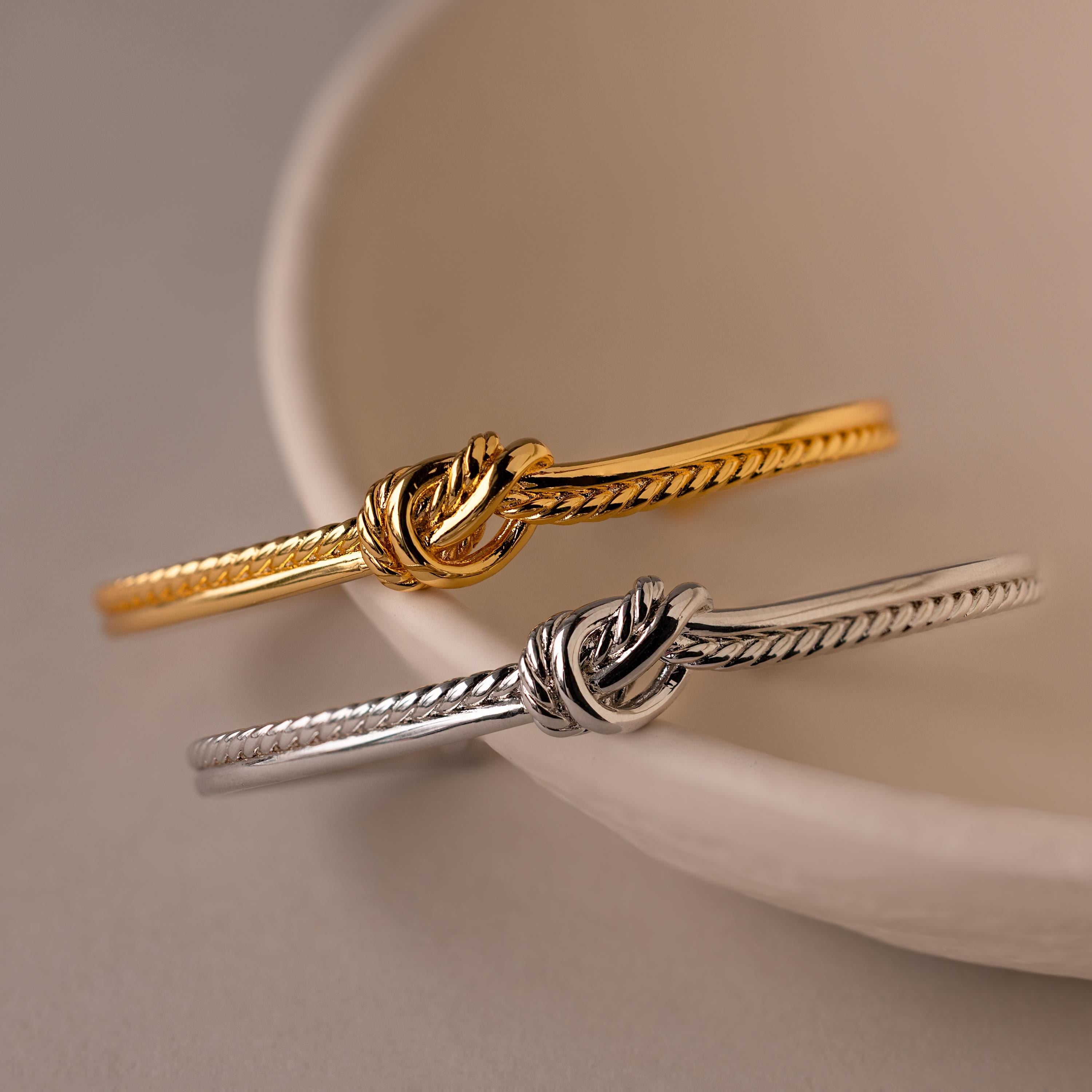Two Duo Knot Bangles, one in gold and one in silver with a delicate rope texture, are displayed on a curved white ceramic dish—making them an elegant gift for any occasion.