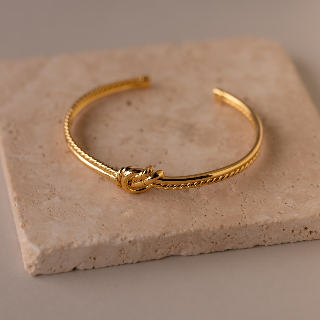 The Duo Knot Bangle, featuring a subtle rope texture and elegant gold finish, is displayed on a beige stone tile—making it a sophisticated gift for any occasion.