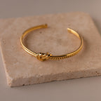 The Duo Knot Bangle, featuring a subtle rope texture and elegant gold finish, is displayed on a beige stone tile—making it a sophisticated gift for any occasion.