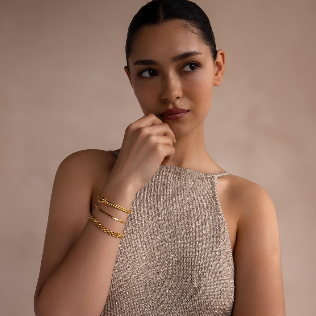 A woman wearing a sleeveless, sparkly beige top and the Duo Knot Bangle with rope texture gazes thoughtfully to the side—a truly elegant gift.