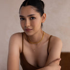 A woman with dark hair in a bun, wearing a brown top and the Strap Necklace—a layered gold piece with interwoven links—gazes at the camera, embodying understated luxury.