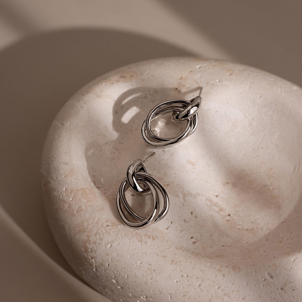 The Bold Duo Knot Earrings are displayed on a smooth, light stone surface with soft shadows that showcase their sculptural and modern design.