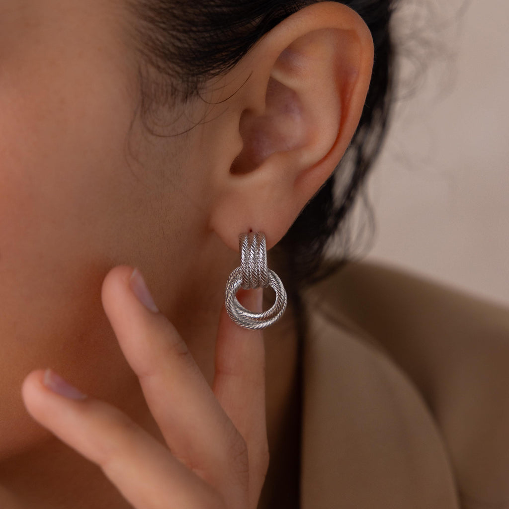A close-up shows someone wearing Ribbed Duo Knot Earrings, gently touching their earlobe—these earrings are a bold, modern accent for any outfit.