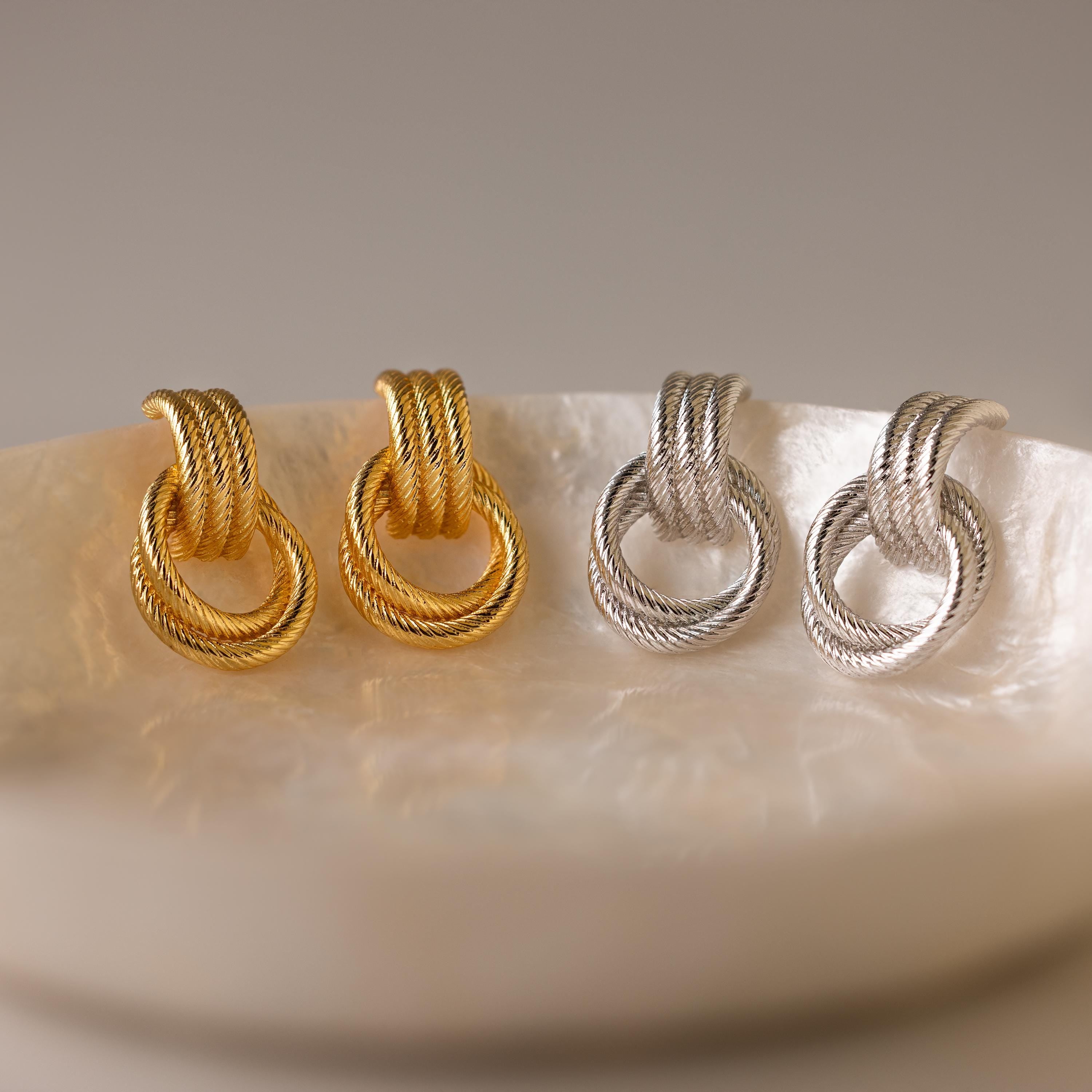 The Ribbed Duo Knot Earrings, in gold and silver, are statement pieces that bring a modern touch to any outfit. Shown on a round iridescent dish, these stylish earrings elevate your look.