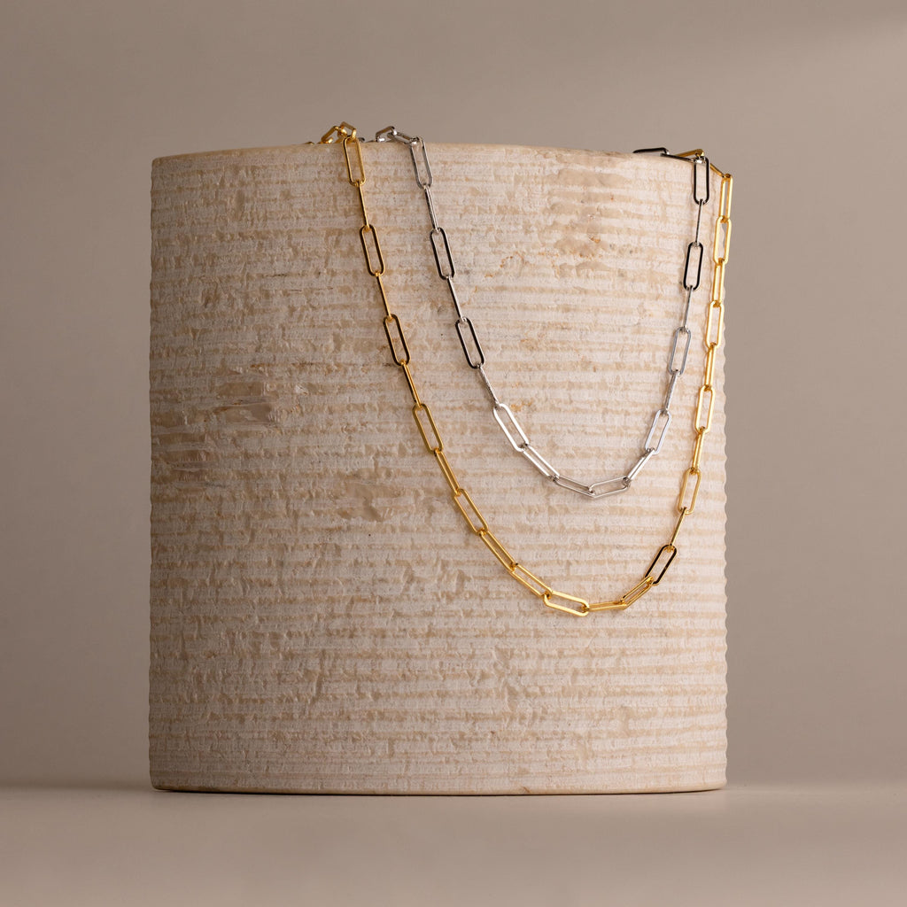 Two chain necklaces, one the Bold Paperclip Chain Necklace in gold and the other in silver, are draped over a textured cylindrical beige object.