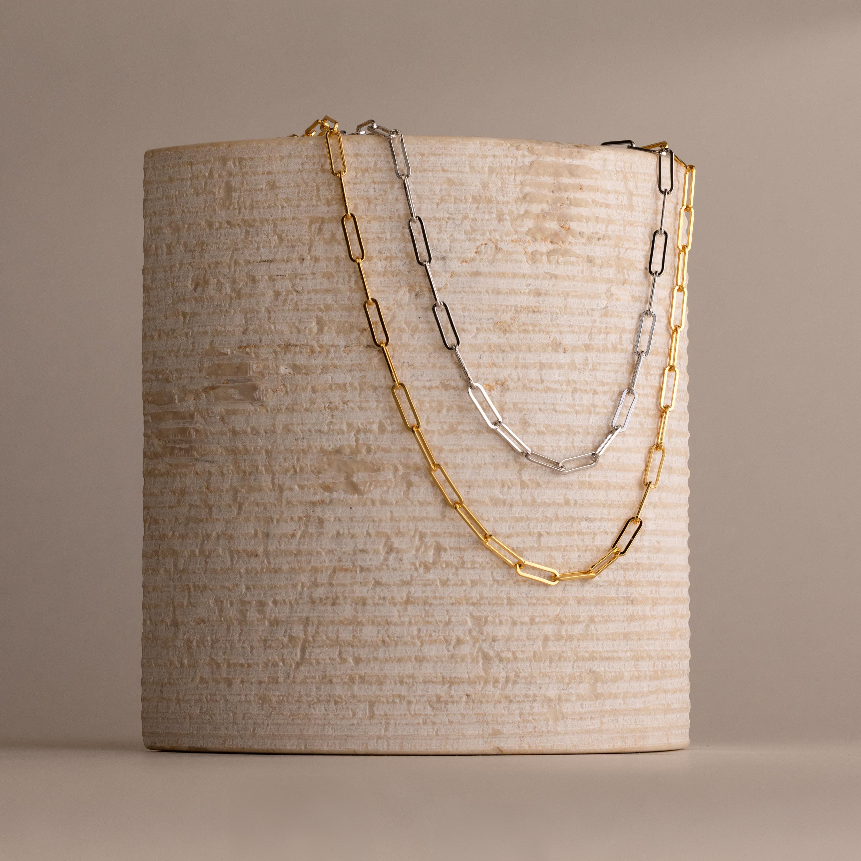 Two chain necklaces, one the Bold Paperclip Chain Necklace in gold and the other in silver, are draped over a textured cylindrical beige object.