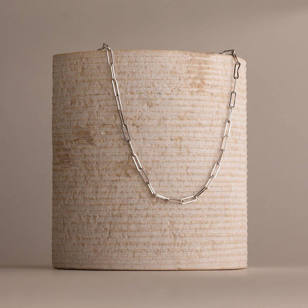 The Bold Paperclip Chain Necklace is elegantly draped over a textured, cylindrical beige stone pedestal.