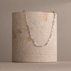 The Bold Paperclip Chain Necklace is elegantly draped over a textured, cylindrical beige stone pedestal.