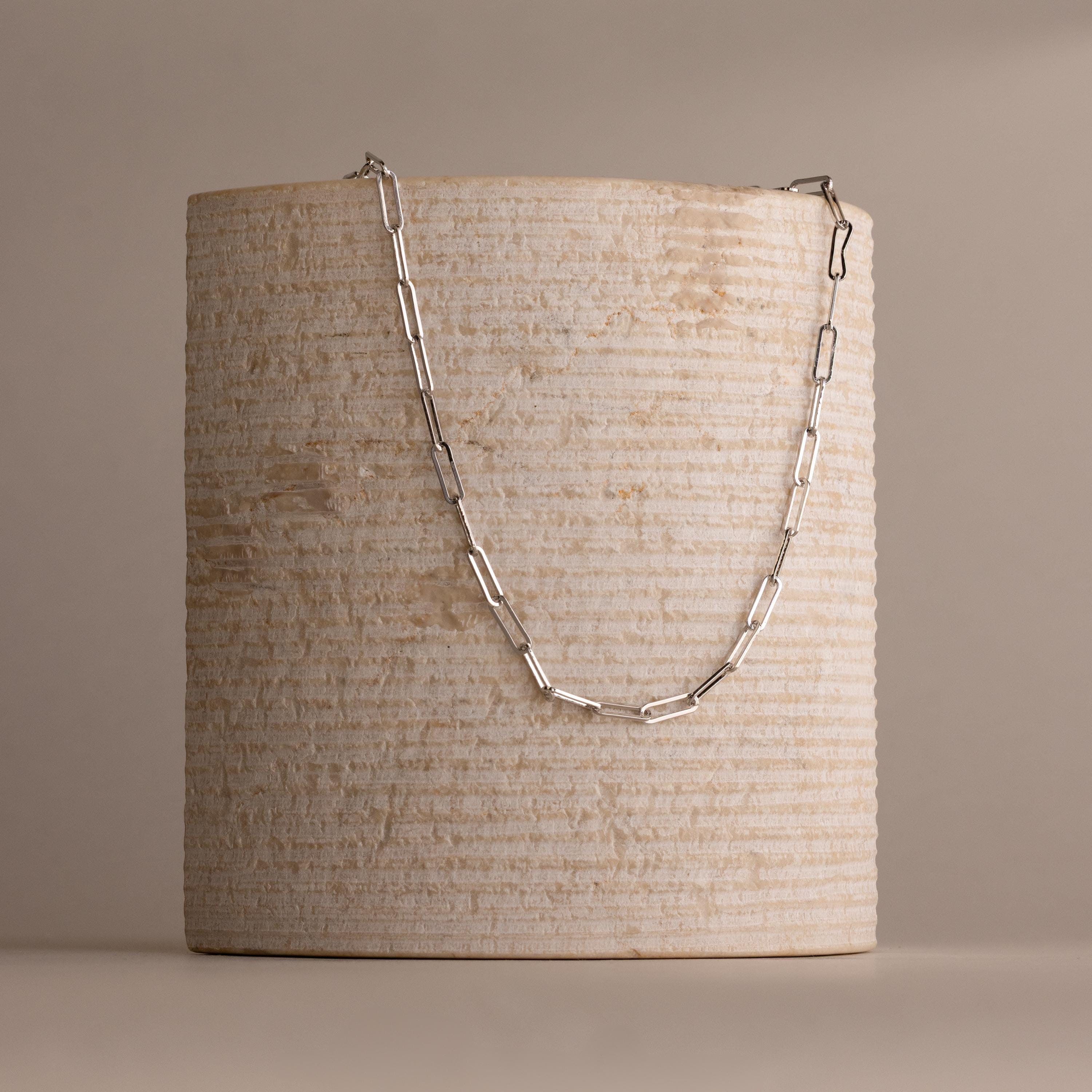 The Bold Paperclip Chain Necklace is elegantly draped over a textured, cylindrical beige stone pedestal.