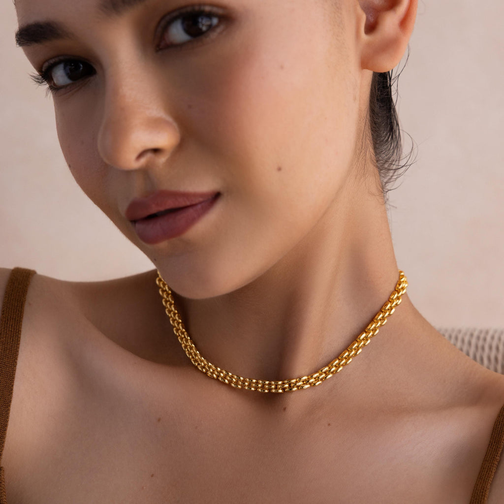 A woman wearing the Strap Necklace—a luxury gold chain with interwoven links—paired with a brown top, gazes at the camera with a neutral expression.