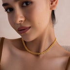 A woman wearing the Strap Necklace—a luxury gold chain with interwoven links—paired with a brown top, gazes at the camera with a neutral expression.