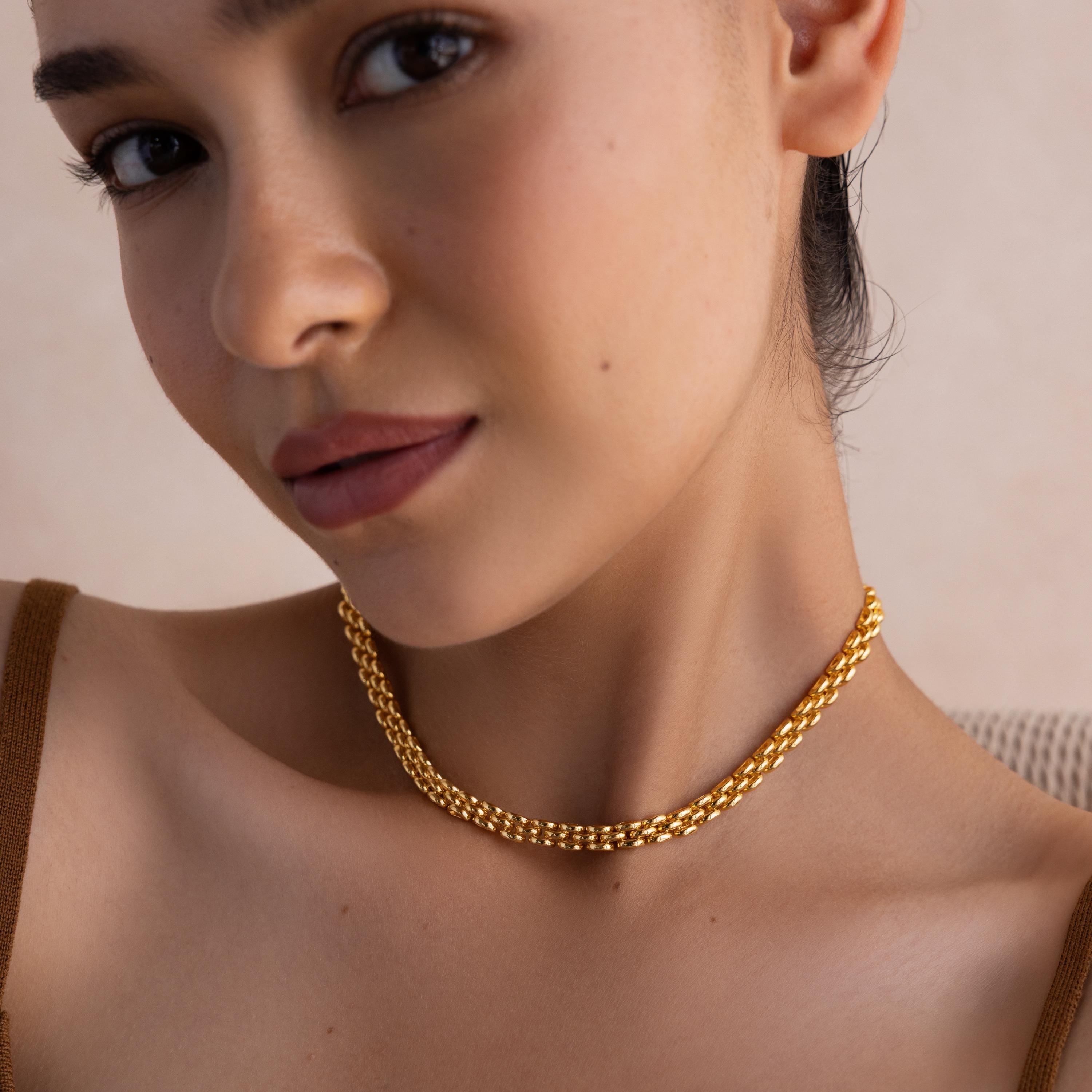A woman wearing the Strap Necklace—a luxury gold chain with interwoven links—paired with a brown top, gazes at the camera with a neutral expression.