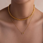 A person wearing the Strap Necklace, a chunky gold choker with interwoven links, layered with a delicate gold chain featuring a small circular pendant for an effortlessly luxurious look.