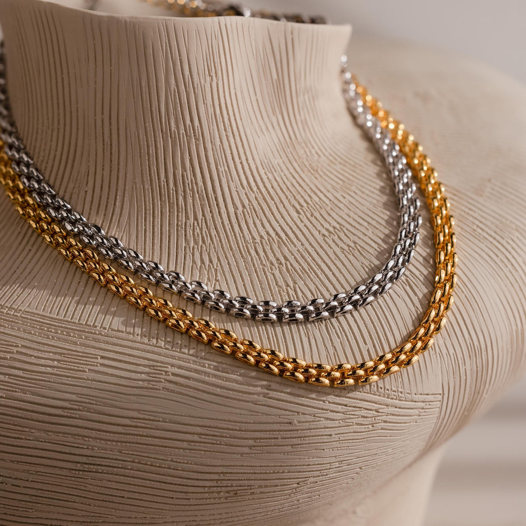 The Strap Necklace, featuring interwoven gold and silver luxury metal chains, is displayed on a textured beige mannequin bust.