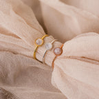Three Dainty Round Moonstone Rings in gold, silver, and rose gold with round moonstones rest on soft beige fabric—a perfect dainty trio for lovers of grounding jewelry.