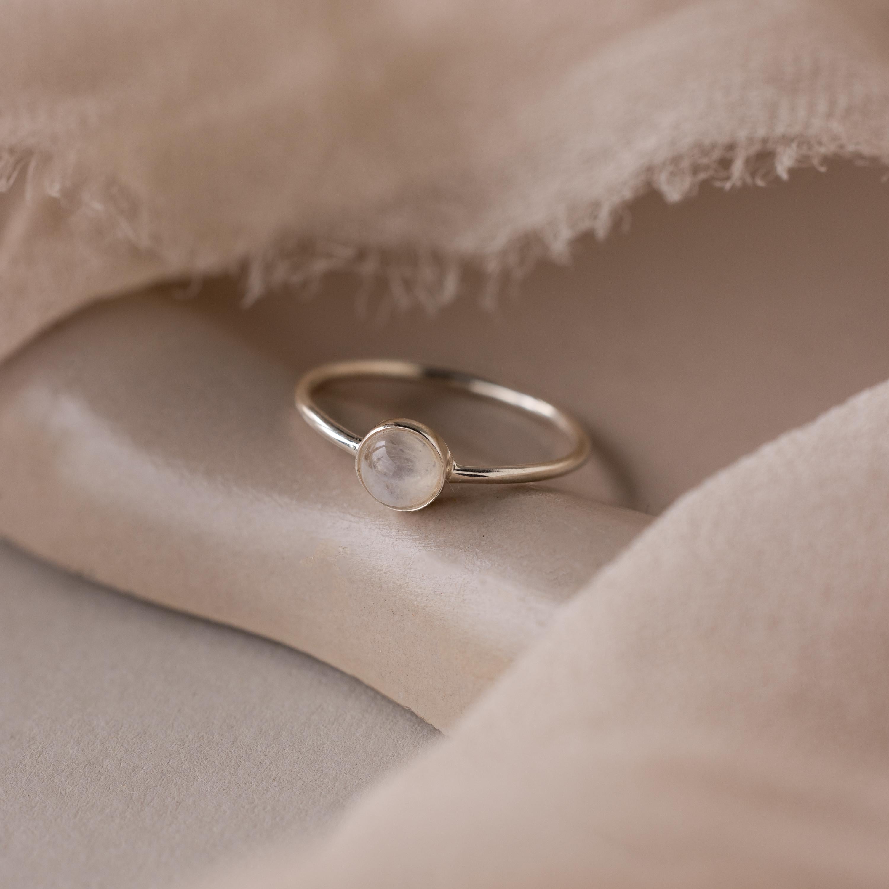The Dainty Round Moonstone Ring features a round, translucent moonstone set on a soft beige fabric background—a perfect, grounding jewelry piece.