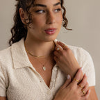 A woman with curly hair wears a cream textured short-sleeve shirt, gold jewelry, and the Pearl Flower Birthstone Necklace with a mother of pearl pendant, looking off to the side with her hands near her face against a plain background.