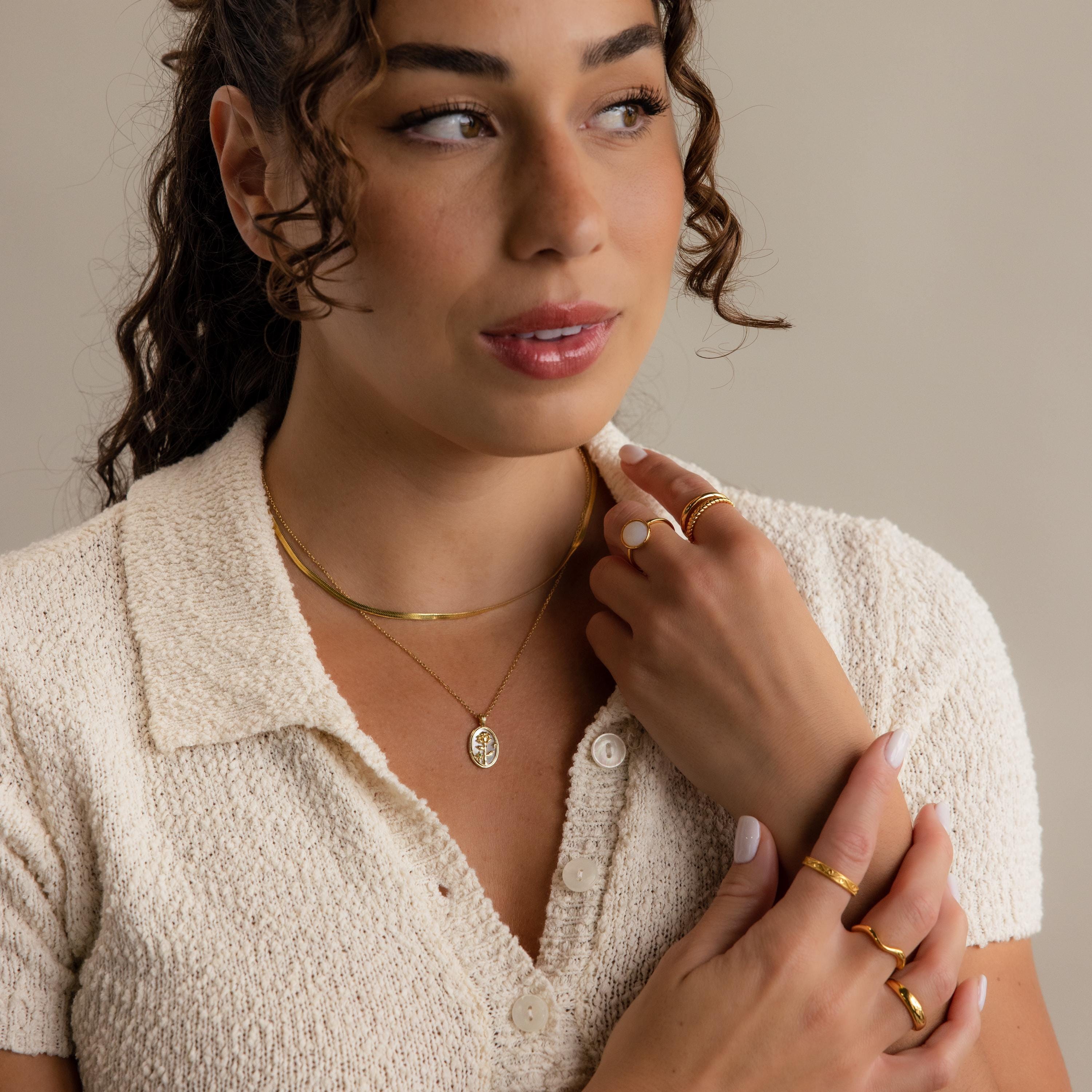 A woman with curly hair wears a cream textured short-sleeve shirt, gold jewelry, and the Pearl Flower Birthstone Necklace with a mother of pearl pendant, looking off to the side with her hands near her face against a plain background.