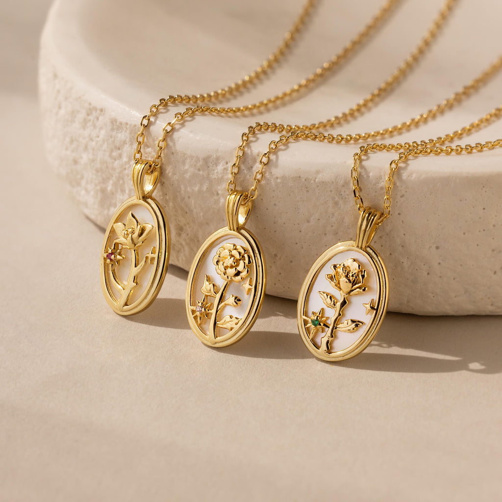 Three Pearl Flower Birthstone Necklaces with gold-colored chains and oval pendants featuring a flower design with a gemstone in the center, displayed on a beige surface.