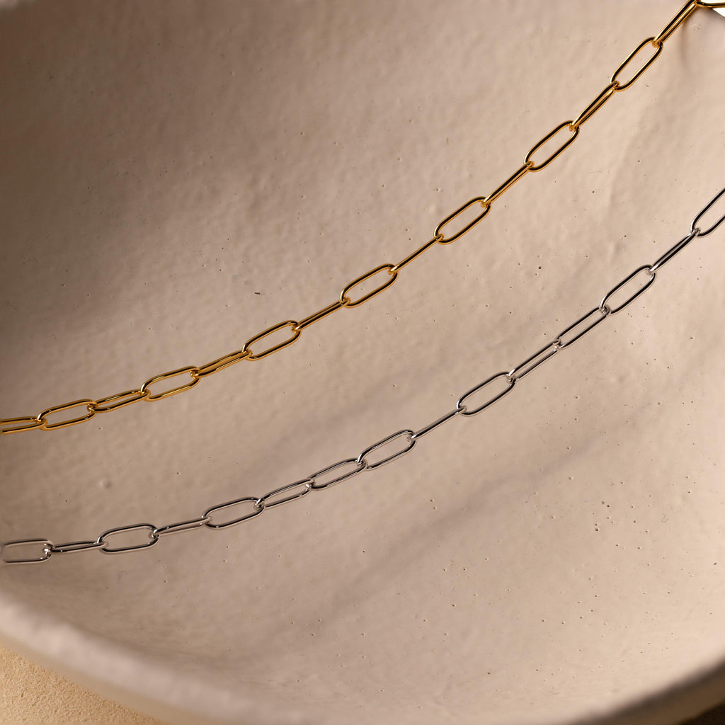 A gold Paperclip Chain Necklace and a delicate silver chain are displayed on an off-white ceramic surface, creating an elegant layered necklace look.