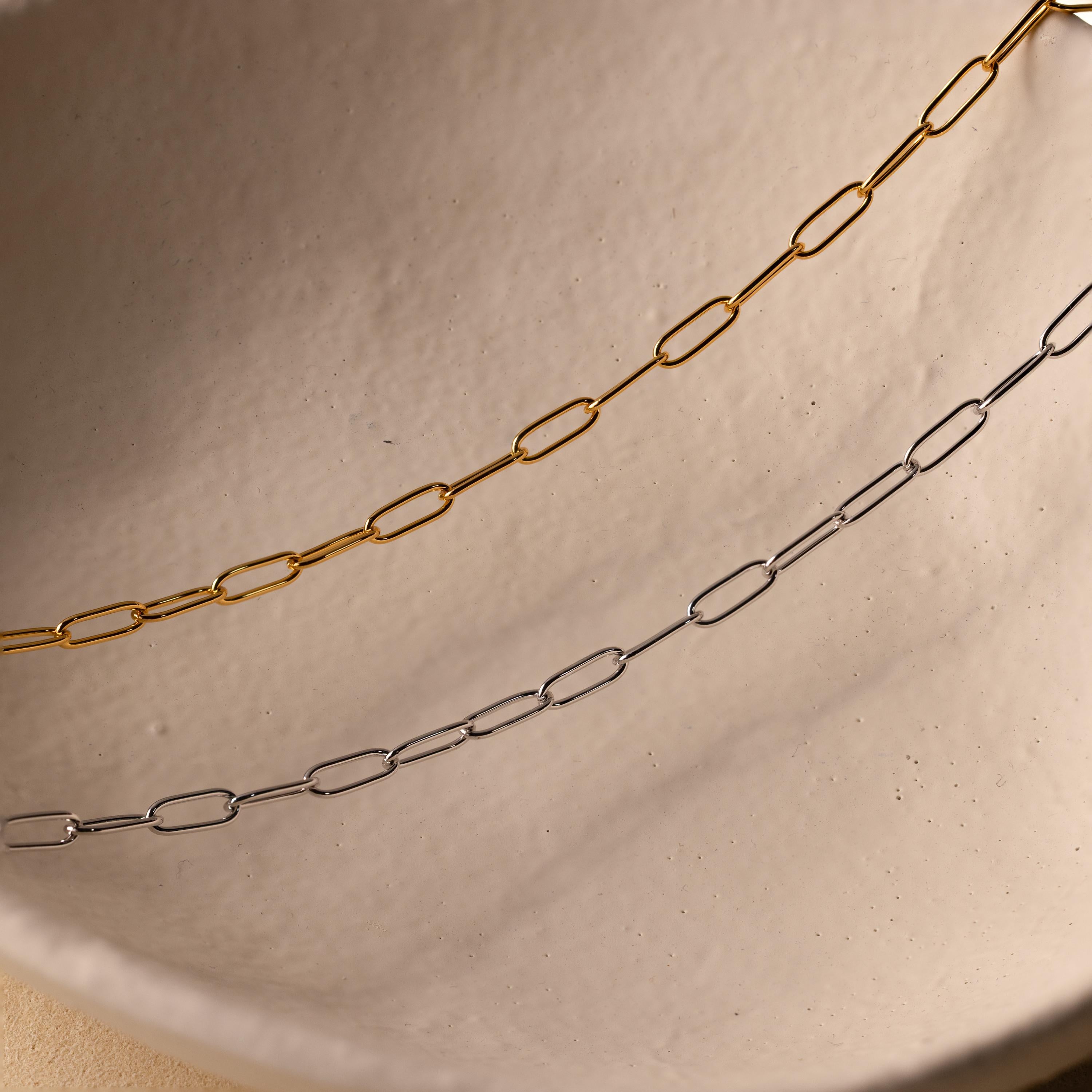 A gold Paperclip Chain Necklace and a delicate silver chain are displayed on an off-white ceramic surface, creating an elegant layered necklace look.