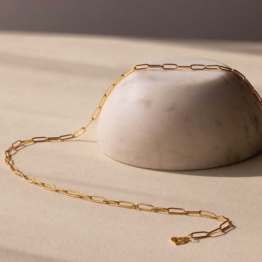 The Paperclip Chain Necklace features elongated gold links, elegantly draped over a smooth, round white stone on a beige surface—ideal for lovers of minimalist jewelry.