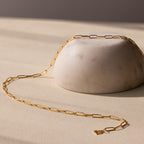 The Paperclip Chain Necklace features elongated gold links, elegantly draped over a smooth, round white stone on a beige surface—ideal for lovers of minimalist jewelry.