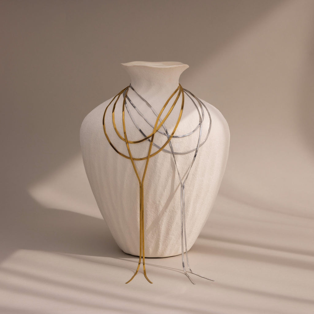 The Long Y Herringbone Chain Necklace, along with thin gold and silver necklaces, drapes over a white vase set against a neutral background.