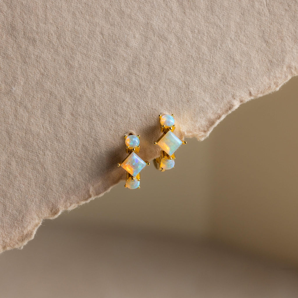 Princess Cut Birthstone Huggies featuring opal-like stones displayed on a textured beige paper background.