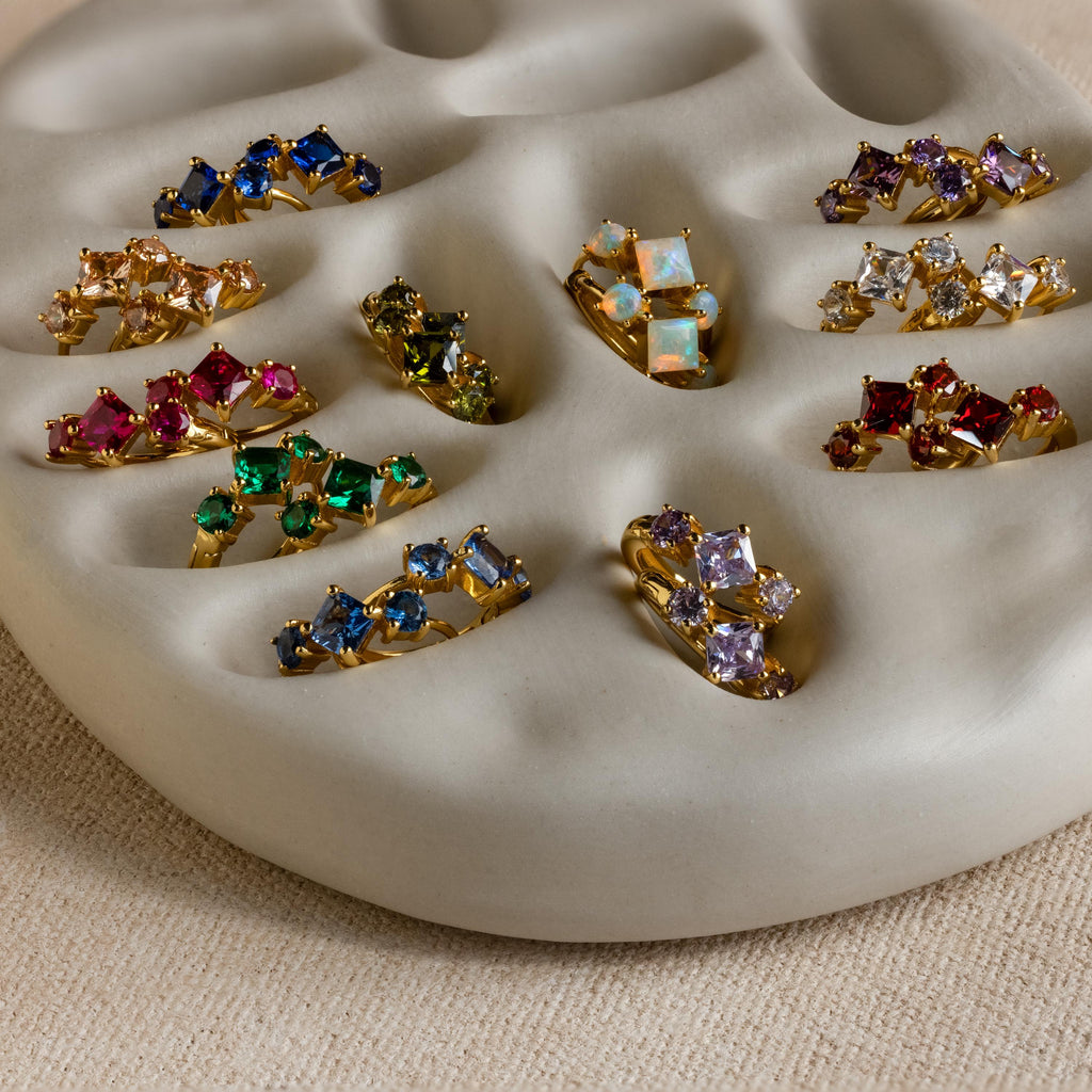 Princess Cut Birthstone Huggies and gold rings with colorful gemstones are displayed on a cream wavy ceramic ring holder, adding a vibrant touch.