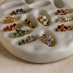 Princess Cut Birthstone Huggies and gold rings with colorful gemstones are displayed on a cream wavy ceramic ring holder, adding a vibrant touch.