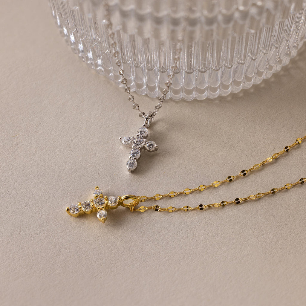 The Diamond Cross Necklace with gemstones and pave CZ diamonds is displayed on a neutral surface near a glass container.