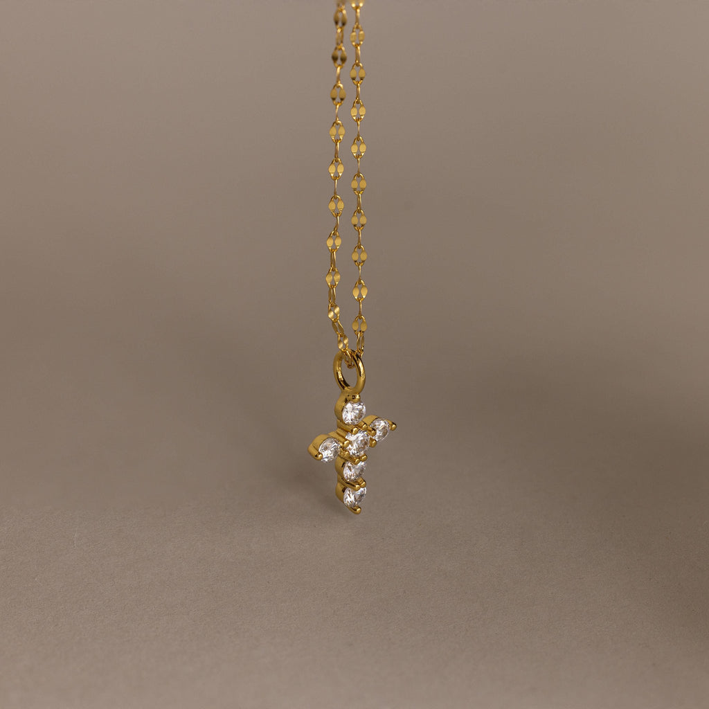 Gold chain with a small, pave CZ diamond cross pendant against a plain beige background, offering a playful twist on classic faith necklace designs.