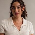 A woman with curly hair smiles softly at the camera, wearing a cream textured blouse and layered gold necklaces, including the Diamond Cross Necklace.