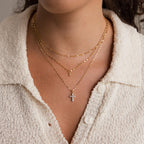 A woman wears three layered gold necklaces, featuring the Diamond Cross Necklace accented with pavé CZ diamonds, over a cream textured button-up top.