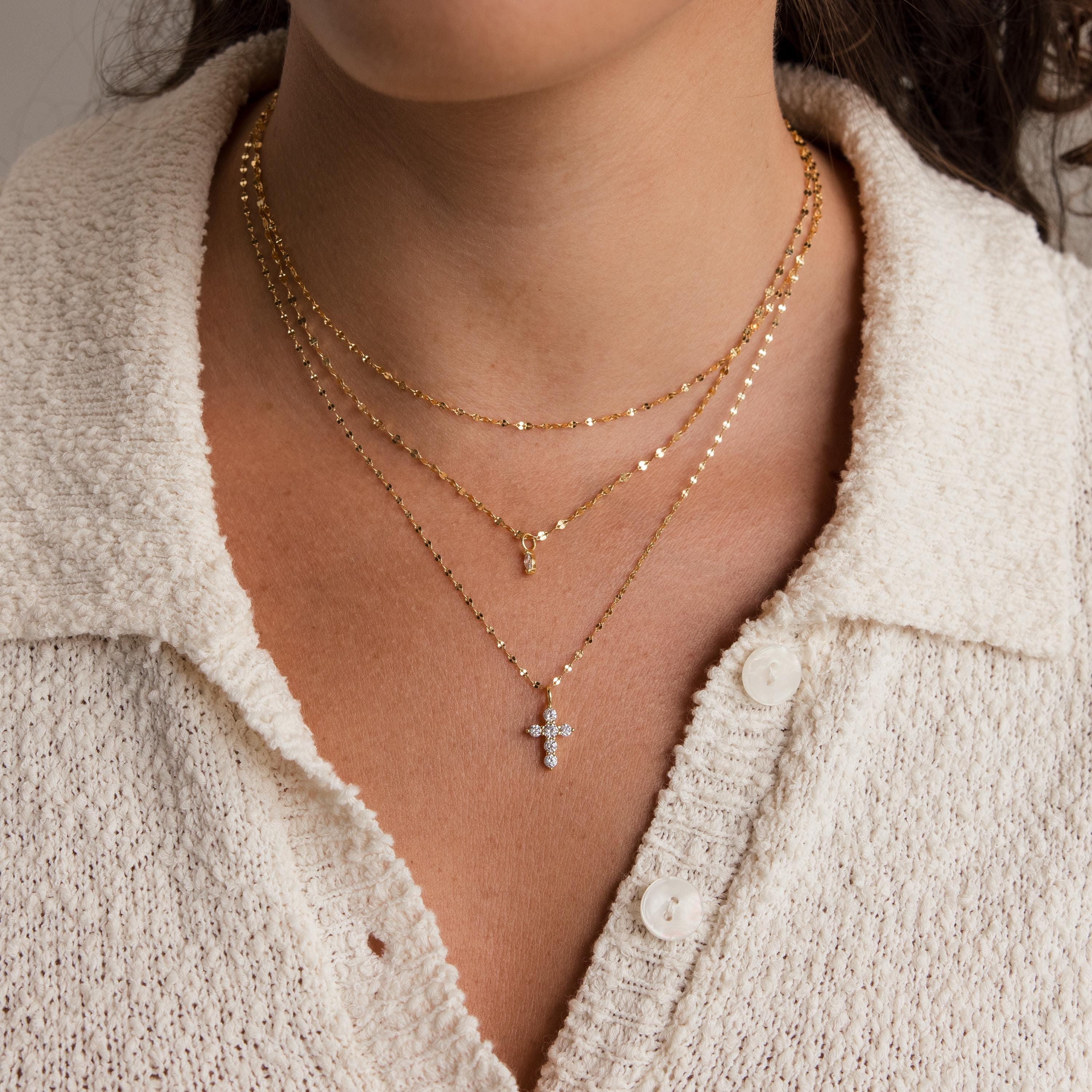 A woman wears three layered gold necklaces, featuring the Diamond Cross Necklace accented with pavé CZ diamonds, over a cream textured button-up top.