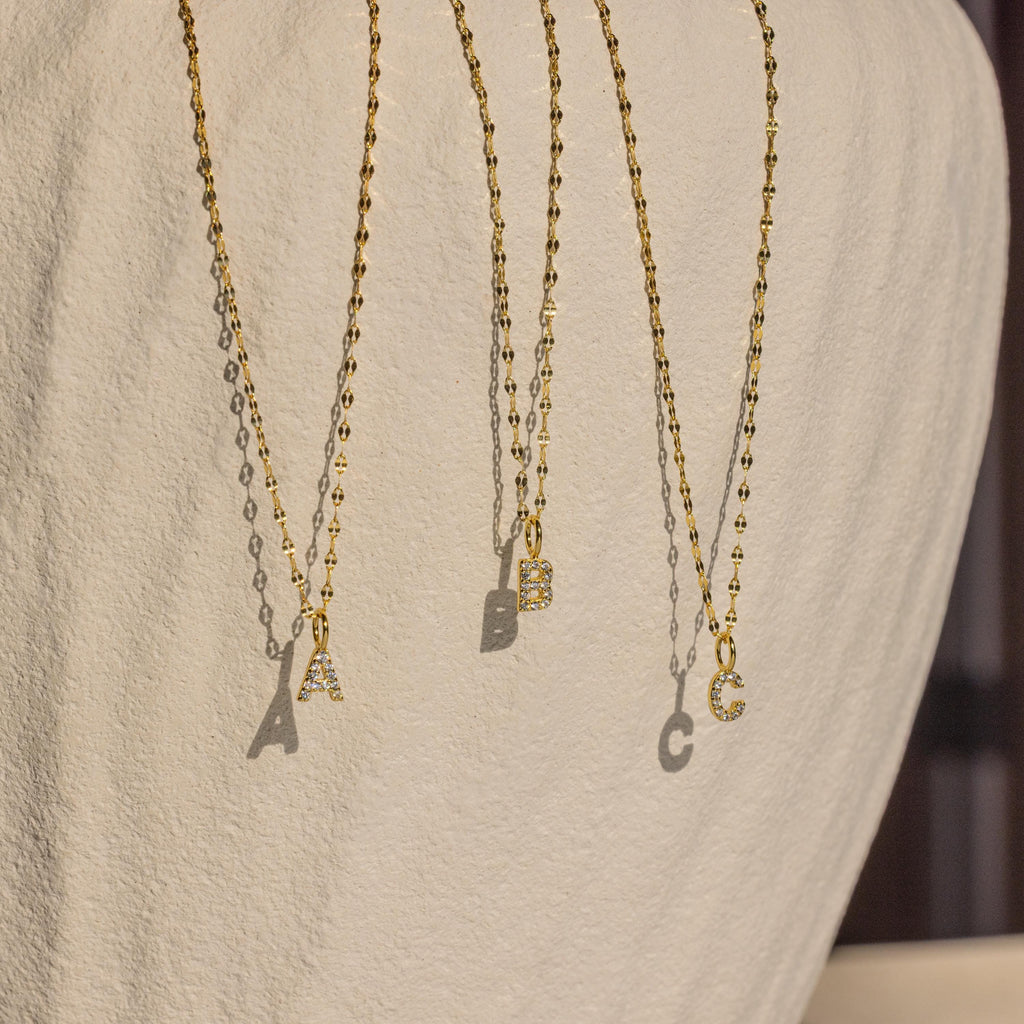 Three Dainty Pave Letter Necklaces, featuring gold chains and pavé CZ diamond-studded A, B, and C pendants, hang elegantly against a textured white background.