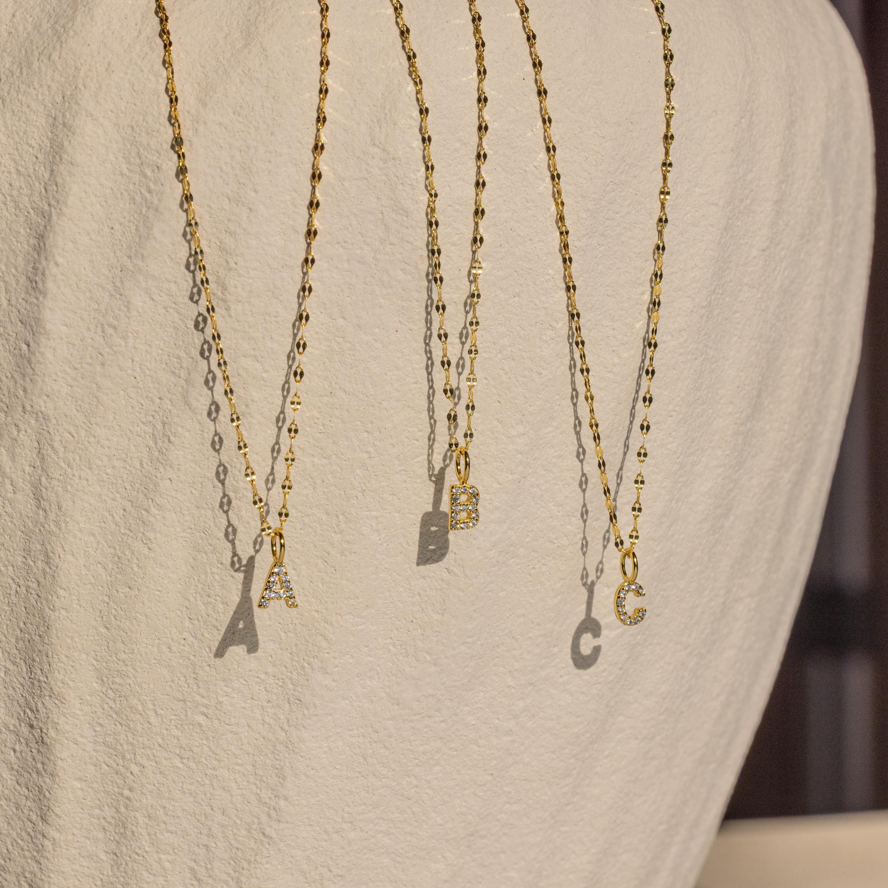 Three Dainty Pave Letter Necklaces, featuring gold chains and pavé CZ diamond-studded A, B, and C pendants, hang elegantly against a textured white background.