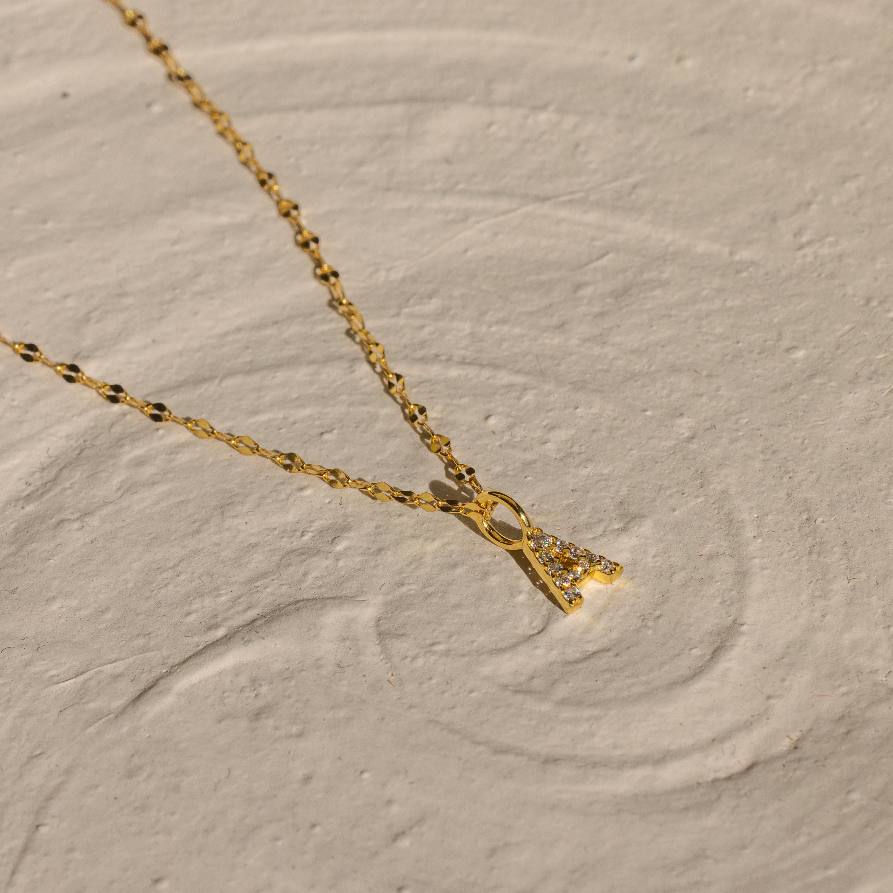The Dainty Pave Letter Necklace in 18K Gold features a sparkling CZ pavé letter pendant, elegantly displayed on a textured beige surface.