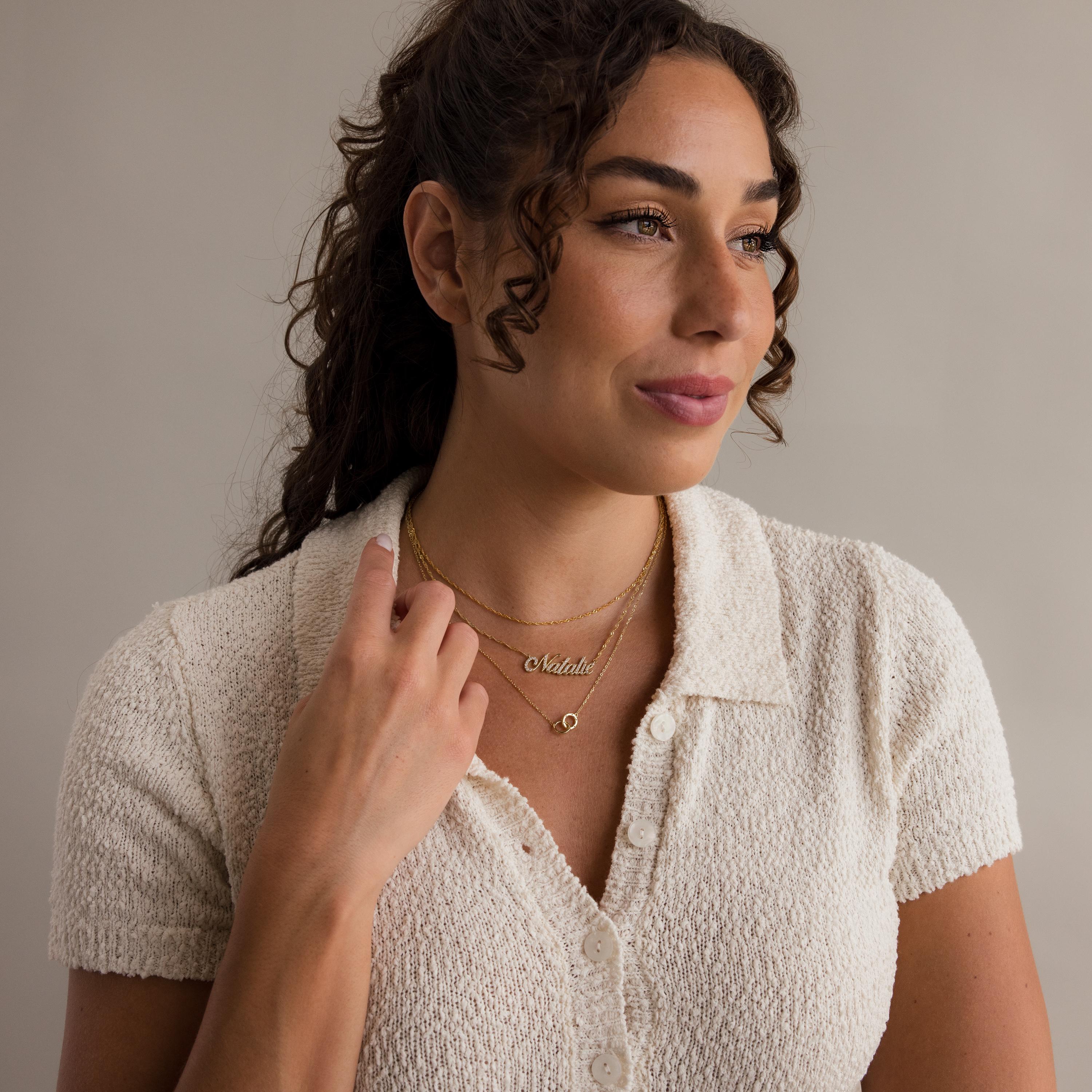 Woman with curly hair wears a cream textured shirt and layered gold necklaces, including the Pave Interlocking Circle Necklace—a symbol of love—while looking to the side.