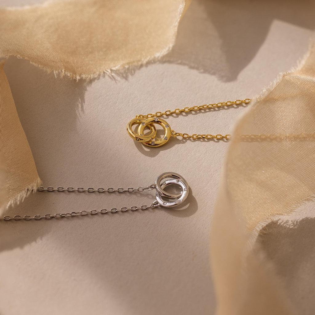 The Pave Interlocking Circle Necklace, featuring gold and silver rings with sparkling CZ diamonds, rests elegantly on a neutral fabric background, beautifully capturing the timeless symbol of love.