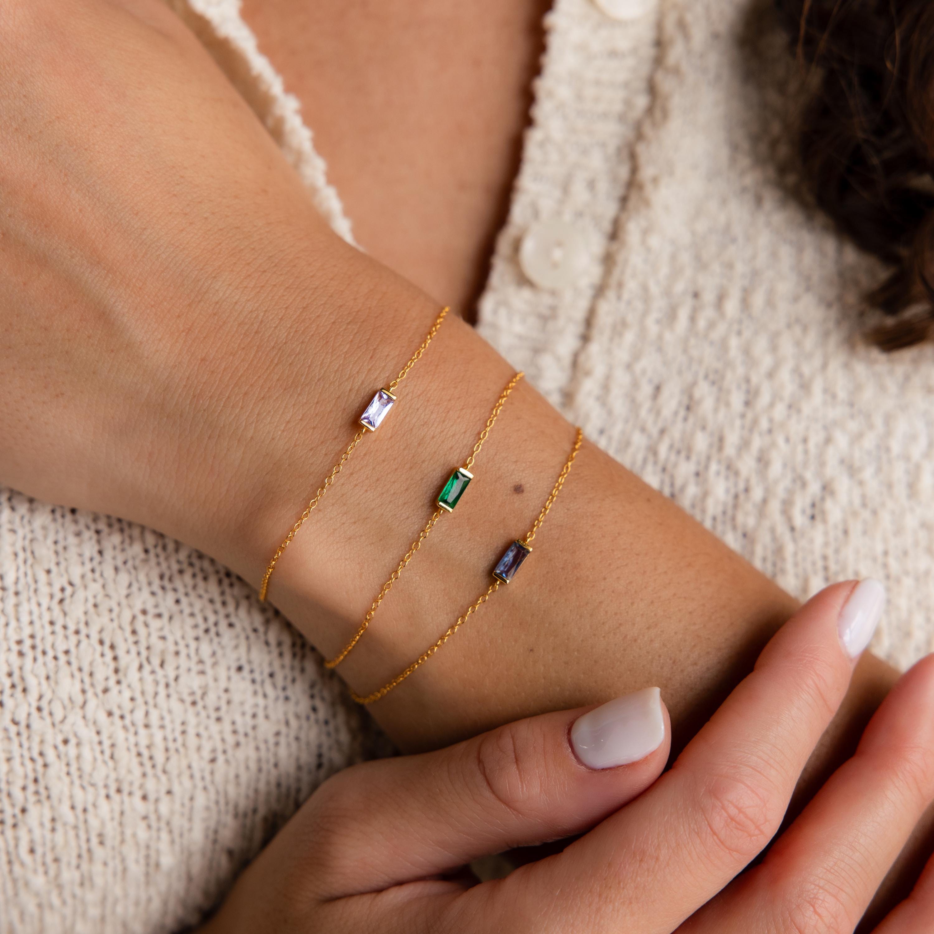 A hand wearing three gold chain bracelets, including the Dainty Baguette Birthstone Bracelet with colorful rectangular gemstones, rests on a cream sweater.