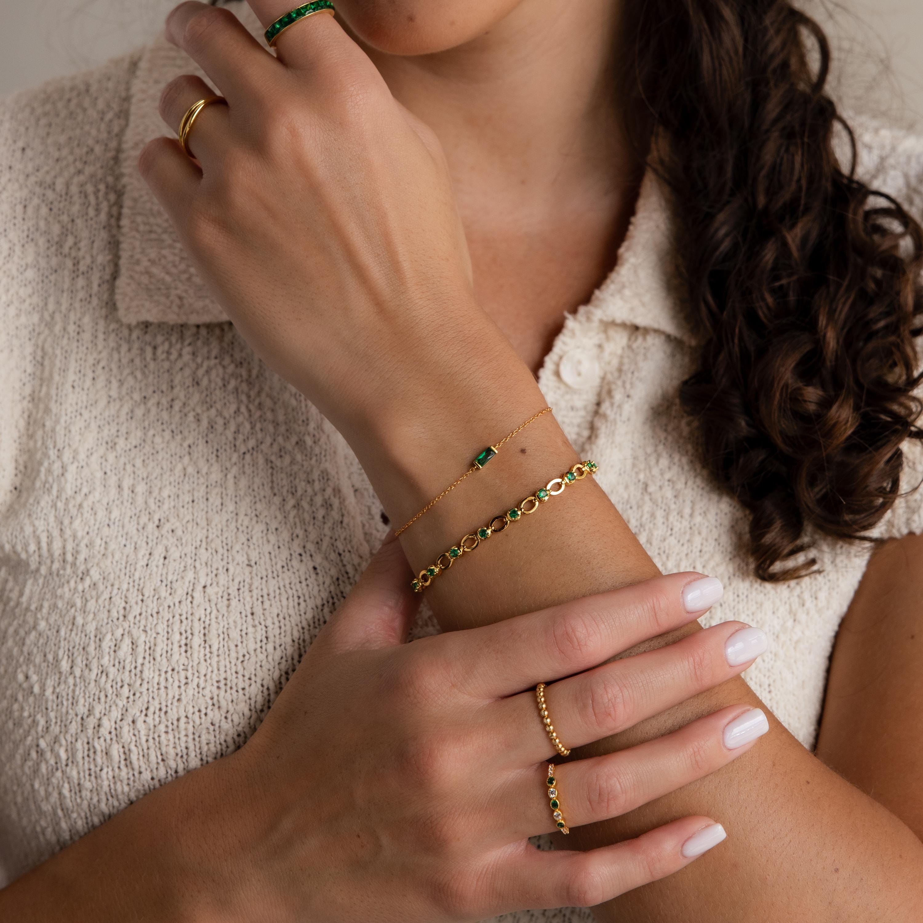 A woman with manicured nails wears gold jewelry, a cream textured top, and the Dainty Baguette Birthstone Bracelet, her hands gracefully crossed.