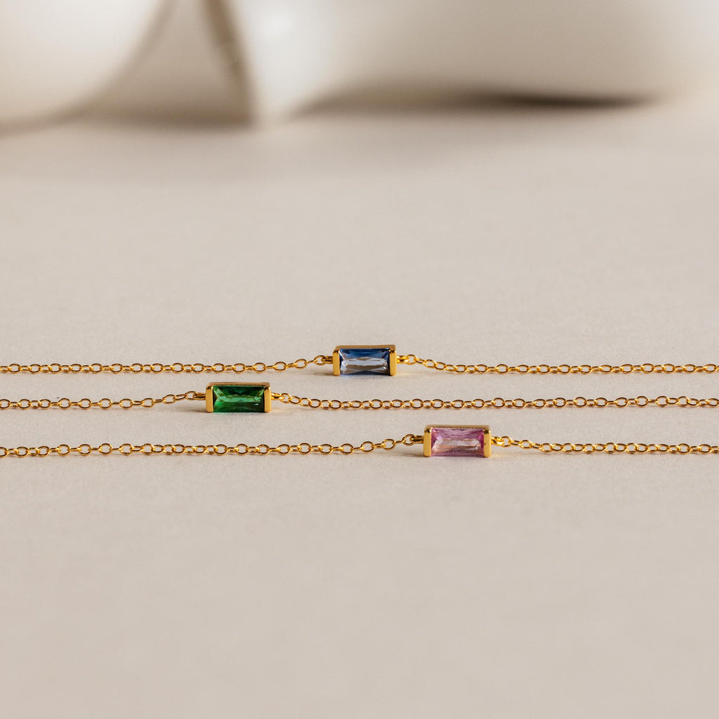 Three gold chain necklaces with rectangular blue, green, and purple gemstones are displayed on a neutral background, reflecting the refined elegance of the Dainty Baguette Birthstone Bracelet.