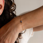 A woman with curly hair wears the Dainty Baguette Birthstone Bracelet, a gold minimalist piece featuring a small blue gemstone on her wrist.
