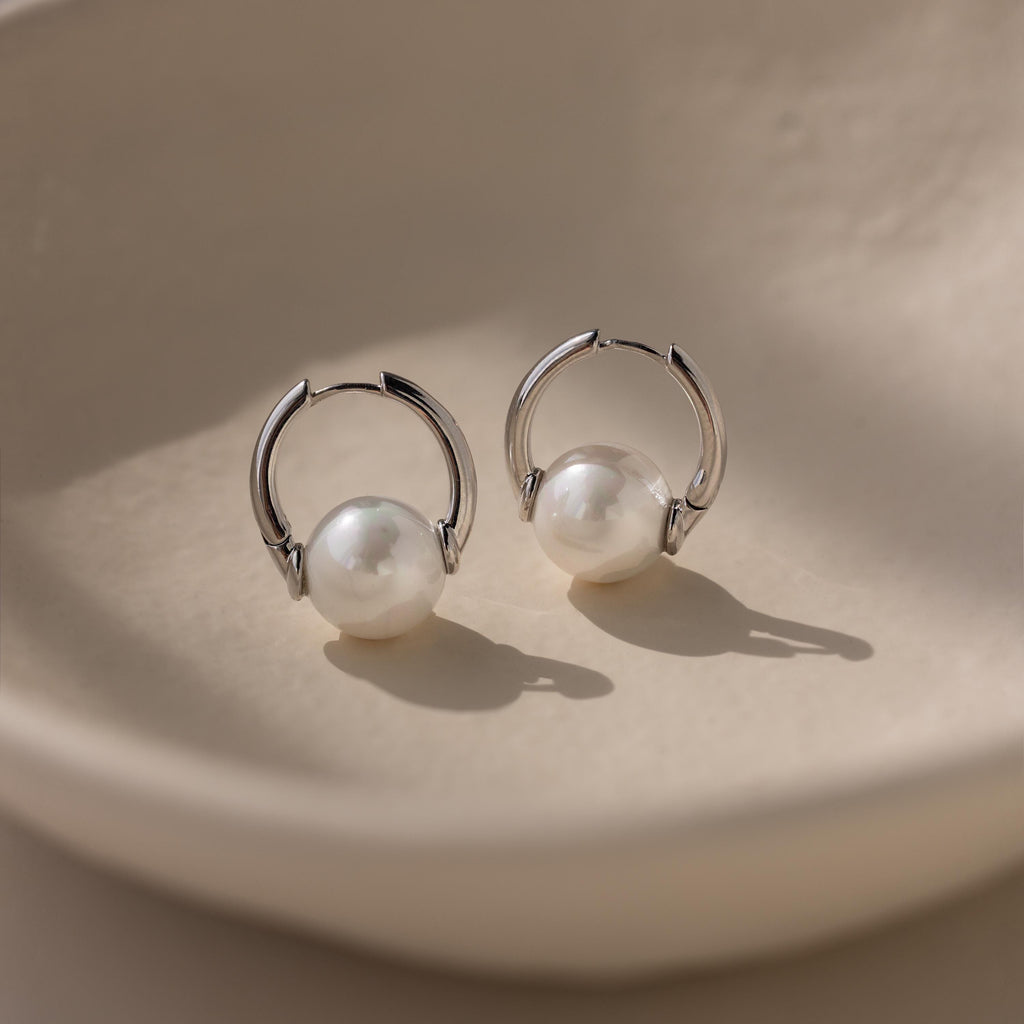 The Bold Round Pearl Hoops, featuring lustrous white pearls, are shown on a smooth cream ceramic dish, making these statement earrings a striking accessory for any occasion.